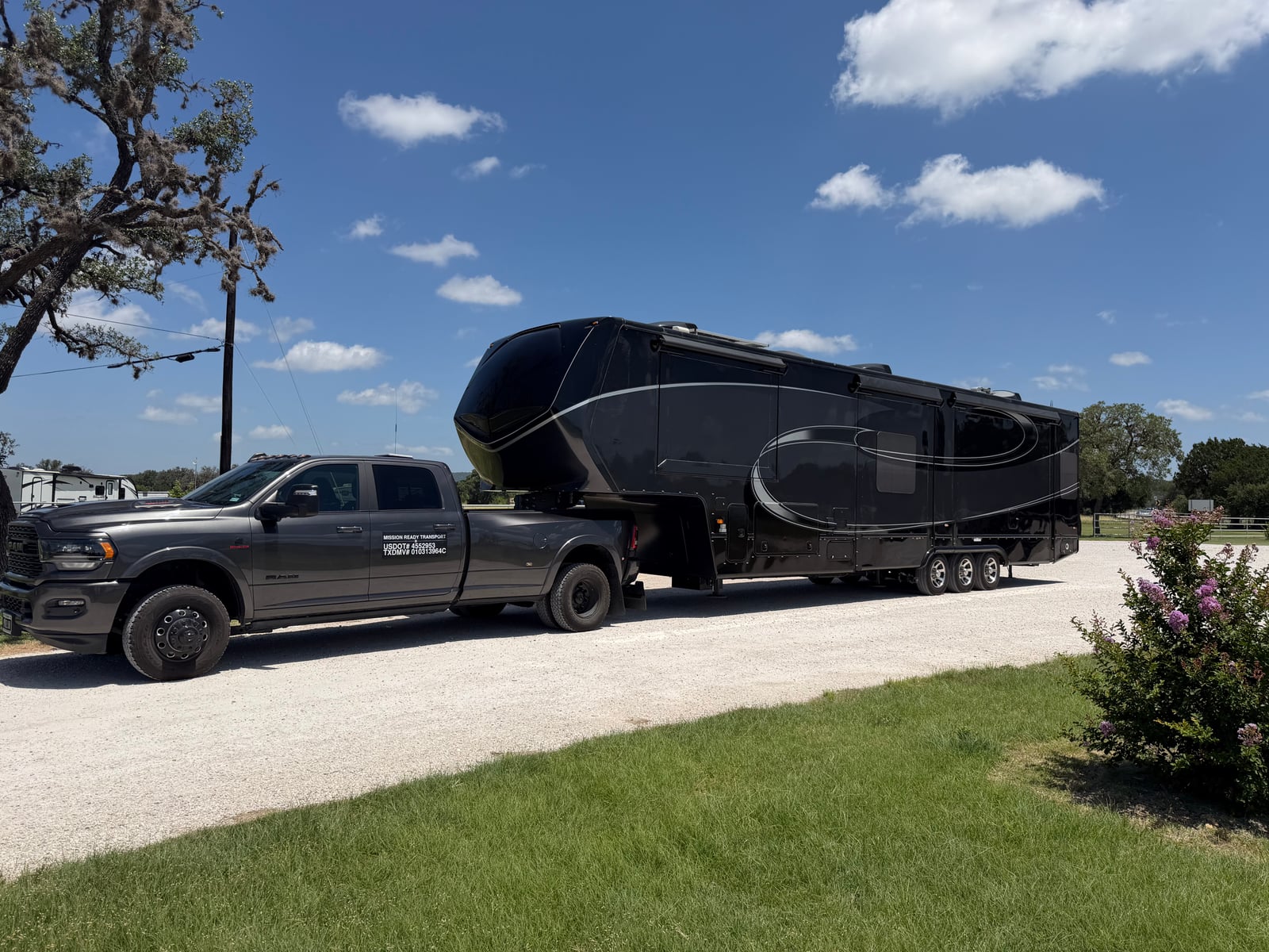 Mission Ready Transport truck towing a large black fifth wheel along a gravel drive